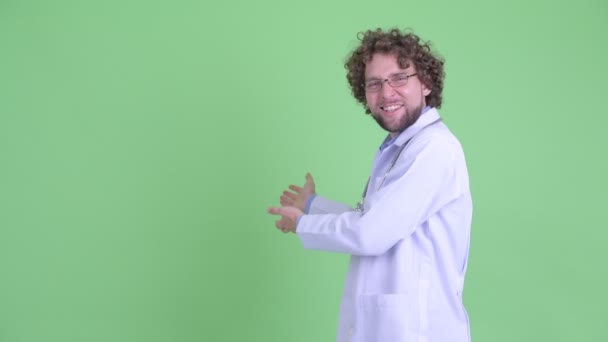 Jovem médico barbudo feliz mostrando algo para trás e recebendo boas notícias — Vídeo de Stock