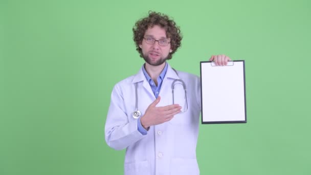 Jovem médico barbudo feliz apresentando com prancheta e dando polegares para cima — Vídeo de Stock