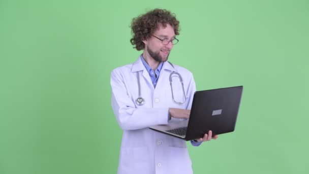 Gelukkige jonge bebaarde man arts denken tijdens het gebruik van laptop — Stockvideo