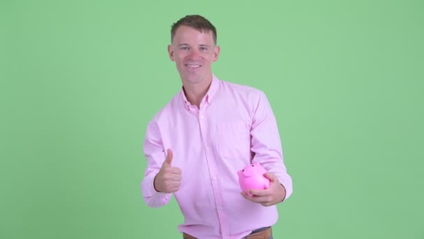 快乐的商人拿着小猪银行，竖起大拇指 — 图库视频影像
