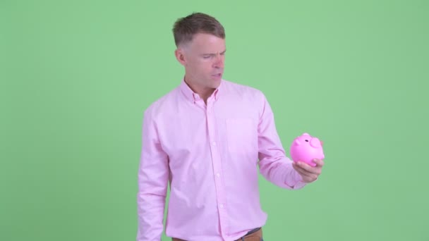 Stressed businessman holding piggy bank and giving thumbs down — Stock Video