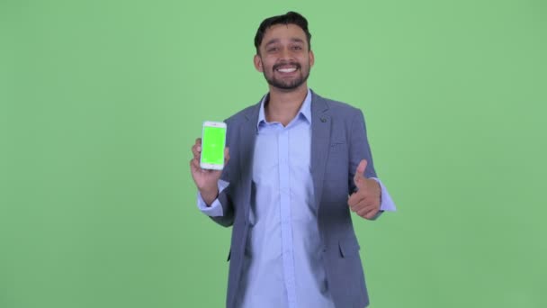 Счастливый молодой бородатый персидский бизнесмен показывает телефон и показывает большие пальцы вверх — стоковое видео