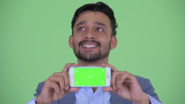 Face of happy young bearded Persian businessman thinking while showing phone — Stock Video