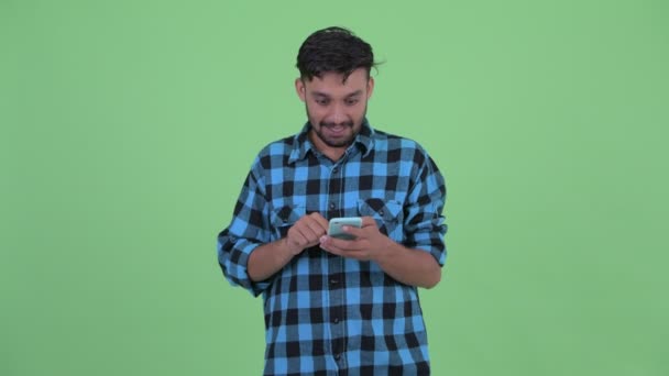 Happy young bearded Persian hipster man using phone and getting good news — Stok Video