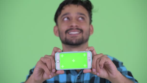 Face of happy young bearded Persian hipster man thinking while showing phone — Stock Video