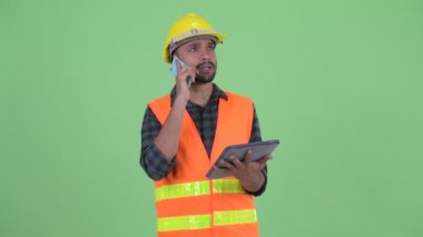 Mutlu genç sakallı Pers adam inşaat işçisi telefon ve dijital tablet kullanarak