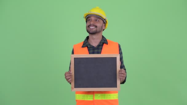 Feliz joven barbudo hombre persa trabajador de la construcción pensando mientras sostiene pizarra — Vídeos de Stock