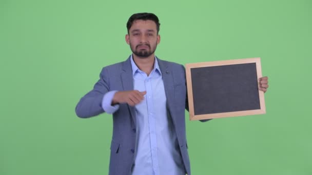 Benadrukte jonge bebaarde Perzische zakenman die Blackboard vasthoudt en duimen neergeeft — Stockvideo