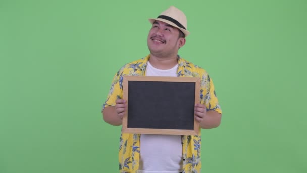 Happy young overweight Asian tourist man thinking while holding blackboard — Stock Video