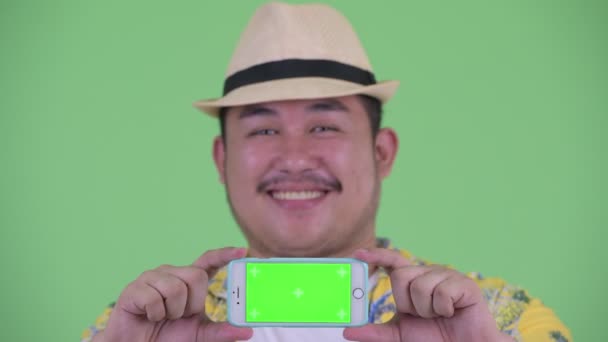 Face of happy young overweight Asian tourist man showing phone — Stock Video