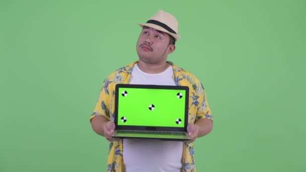 Счастливый молодой толстый азиатский турист думает, показывая ноутбук — стоковое видео