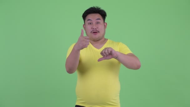 Запутавшийся молодой толстый азиат выбирает между большим и большим пальцами вниз — стоковое видео