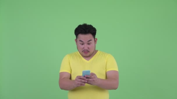 Πρόσωπο του ευτυχισμένο νεαρό υπέρβαρο ασιατικό άτομο χρησιμοποιώντας το τηλέφωνο και αναζητούν έκπληκτος — Αρχείο Βίντεο