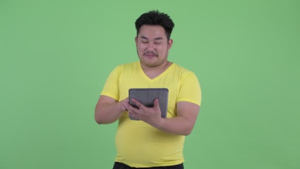 Feliz joven sobrepeso asiático hombre pensando mientras se utiliza la tableta digital — Vídeos de Stock