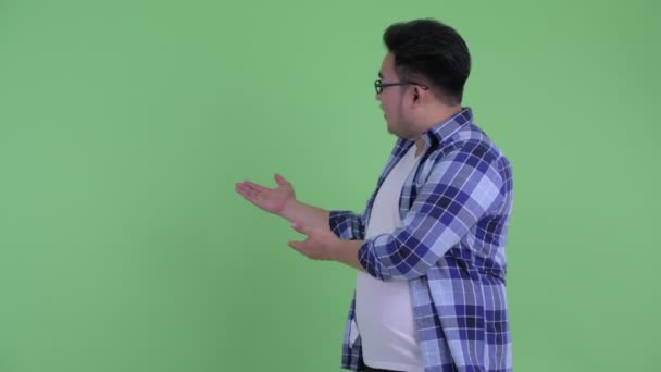 Feliz jovem acima do peso asiático hipster homem falando enquanto mostrando para trás — Vídeo de Stock