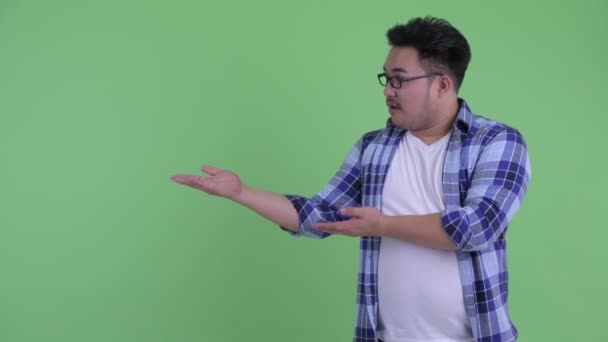 Happy young overweight Asian hipster man talking while showing something — Stock Video