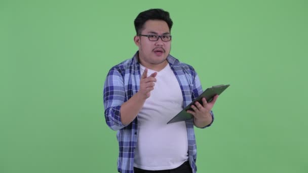 Happy young overweight Asian hipster man talking while holding clipboard — Stock Video