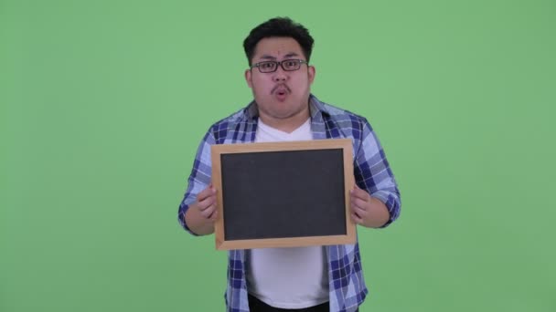 Mutlu genç kilolu Asya hipster adam kara tahta tutan ve şaşırmış görünümlü — Stok video