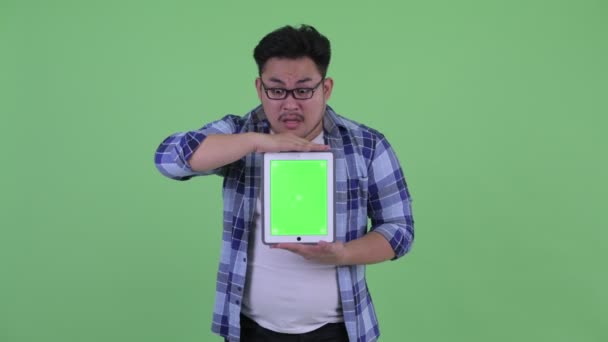 Happy young overweight Asian hipster man showing digital tablet and looking surprised — Stock Video