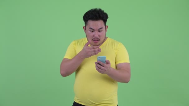 幸せな若い太りすぎのアジアの男性は、携帯電話を使用して、驚いたように見えます — ストック動画