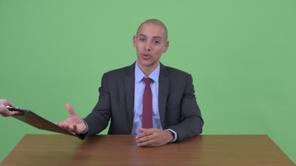 Glücklich glatzköpfige multiethnische Geschäftsmann spricht, während er Klemmbrett hinter Schreibtisch hält — Stockvideo