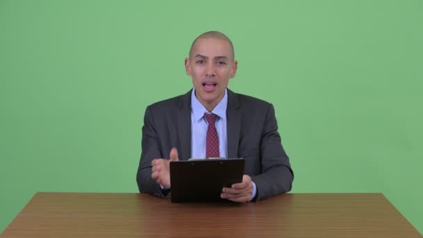 Gestresster multiethnischer Geschäftsmann mit Glatze zeigt Klemmbrett und drückt Daumen nach unten hinter Schreibtisch — Stockvideo