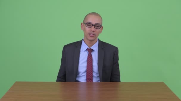 Estresado hombre de negocios calvo multiétnico hablando mientras sostiene el portapapeles detrás del escritorio — Vídeos de Stock