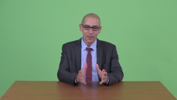 Happy bald multi ethnic businessman presenting while holding clipboard behind desk — Stock Video