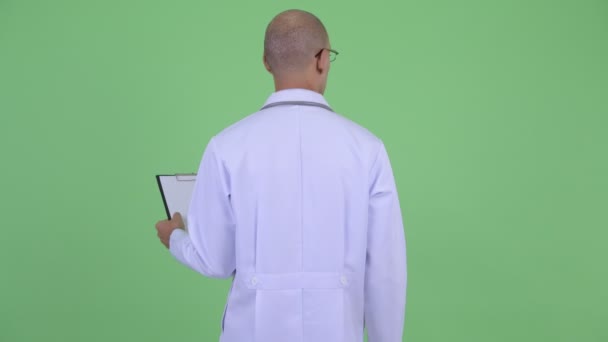 Vista trasera del calvo hombre multiétnico médico pensando y leyendo en el portapapeles — Vídeo de stock