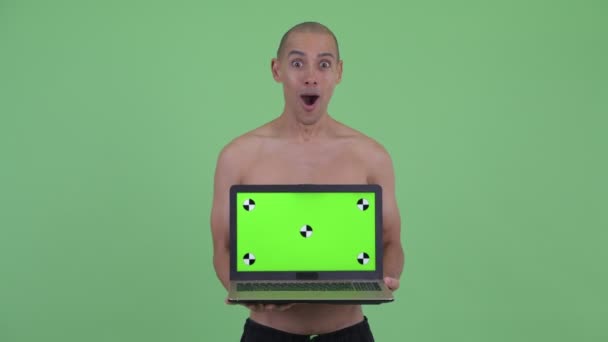 Щасливий лисий багатоетнічний без сорочки чоловік показує ноутбук і виглядає здивовано — стокове відео