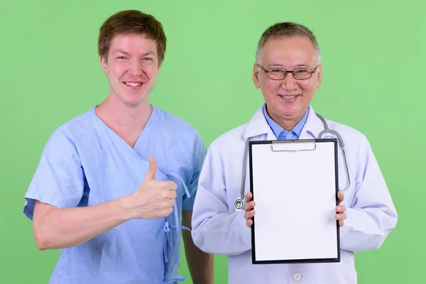 Счастливый зрелый японский врач мужчина со скандинавским пациентом показывая планшет и показывая большие пальцы вверх — стоковое фото