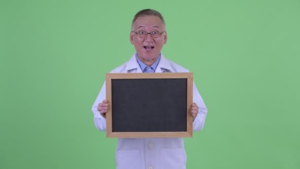 Feliz madura japonesa hombre médico buscando sorprendido mientras sostiene pizarra — Vídeo de stock