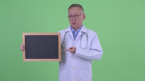 Stressed mature Japanese man doctor holding blackboard and giving thumbs down — Stock Video