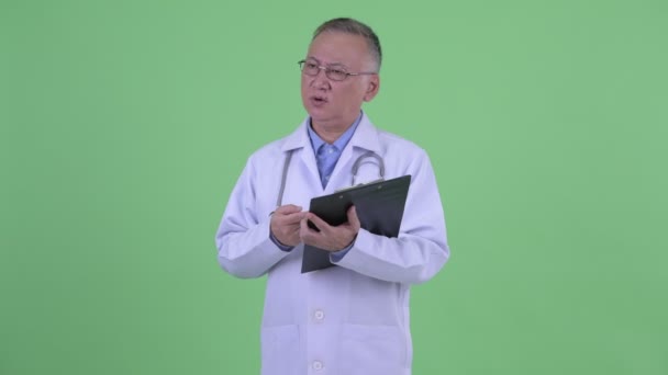 Feliz madura japonesa hombre médico explicando mientras sostiene portapapeles — Vídeos de Stock