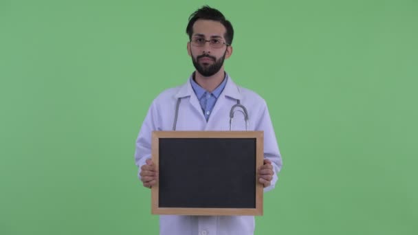 Happy young bearded Persian man doctor talking while holding blackboard — Stock Video