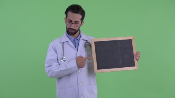 Stressed young bearded Persian man doctor holding blackboard and giving thumbs down — Stock Video