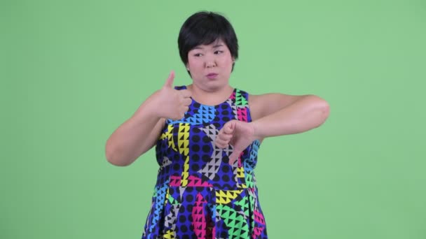 Confused young overweight Asian woman choosing between thumbs up and thumbs down — Stock Video