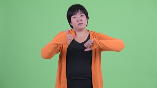 Запутавшаяся молодая толстая азиатка выбирает между большим и большим пальцами вниз — стоковое видео