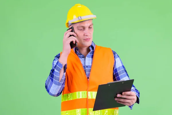 Young man construction worker talking on the phone while holding clipboard — Stock Photo, Image