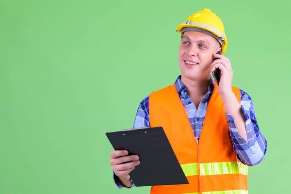 Happy young man construction worker talking on the phone while holding clipboard — Stock Photo, Image