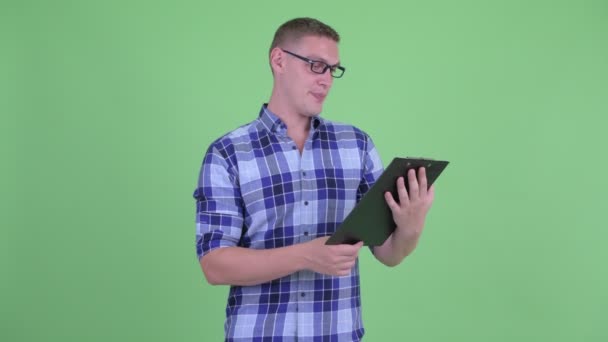 Feliz joven hipster hombre hablando mientras lee en el portapapeles — Vídeos de Stock