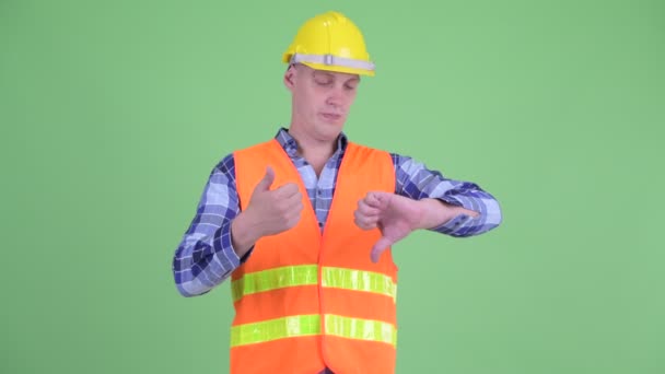 Запутавшийся молодой человек строитель выбирает между большим и большим пальцами вниз — стоковое видео