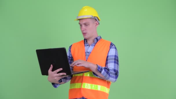 Gelukkige jonge man bouwvakker denken tijdens het gebruik van laptop — Stockvideo