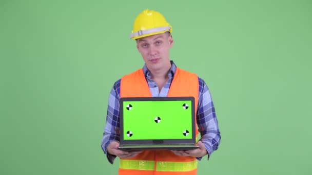 Serious joven trabajador de la construcción mostrando portátil y asintiendo con la cabeza no — Vídeos de Stock