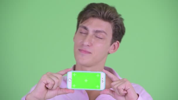 Gesicht eines glücklichen jungen gutaussehenden Geschäftsmannes, der beim Vorzeigen seines Telefons denkt — Stockvideo