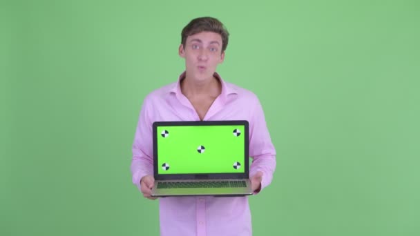 Jovem empresário bonito feliz mostrando laptop e olhando surpreso — Vídeo de Stock
