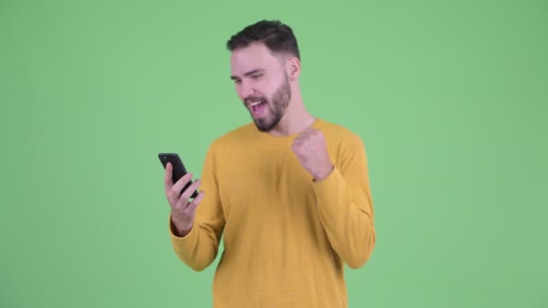 Feliz joven guapo barbudo hombre usando el teléfono y recibiendo buenas noticias — Vídeo de stock