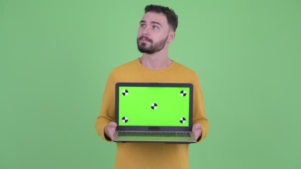 Felice giovane bell'uomo barbuto pensando mentre mostra il computer portatile — Video Stock
