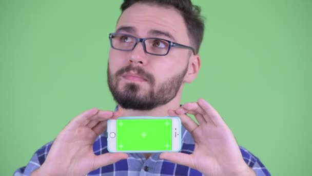 Face of happy young bearded hipster man thinking while showing phone — Stock Video