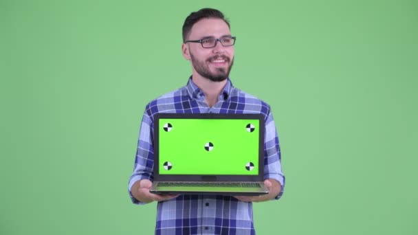 Ευτυχισμένος νεαρός γενειοφόρος hipster άνθρωπος σκέφτεται, ενώ δείχνει φορητό υπολογιστή — Αρχείο Βίντεο